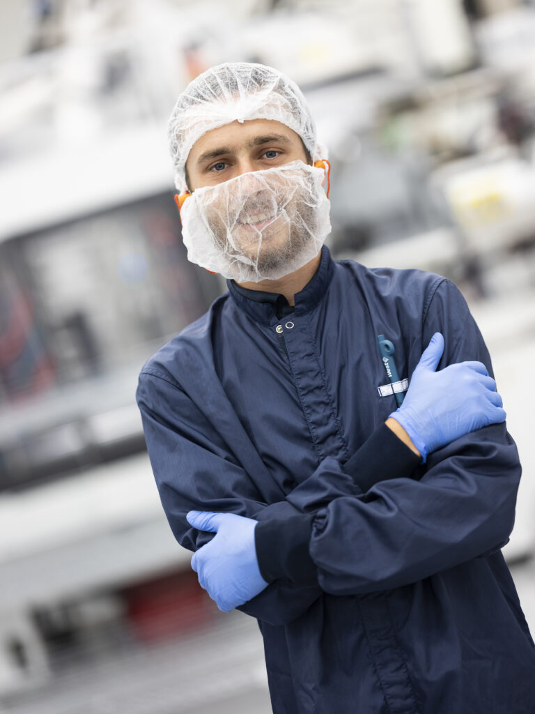 A Happy male employee working at a pharmaceutical factory and looking at the camera smiling wearing protective clothing, hair and beard coverings.