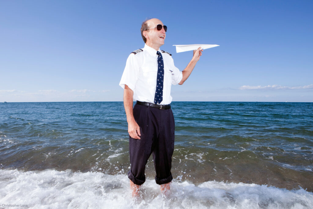 Global Assignments agency New York, New Jersey-based photographer Christopher Lane People Portrait Documentary Reportage Corporate Environmental Social Responsibility Social Enterprise Shared Values Lifestyle Photographer Man dressed as Pilot with a white shirt, applets, trousers rolled up, stands in the Sea Ocean with Paper a Plane
