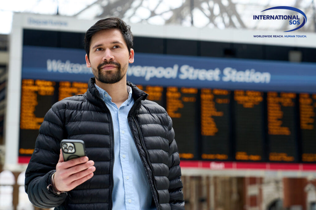 Handheld shot of bearded young man holding mobile phone with train information board behind him. Confident young male professional standing in front of departure board. 