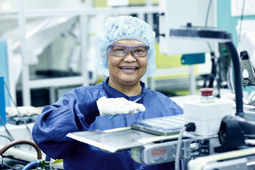 A smiling Asian lady working in a pharmaceutical factory wearing a hair net and blue protective overalls.